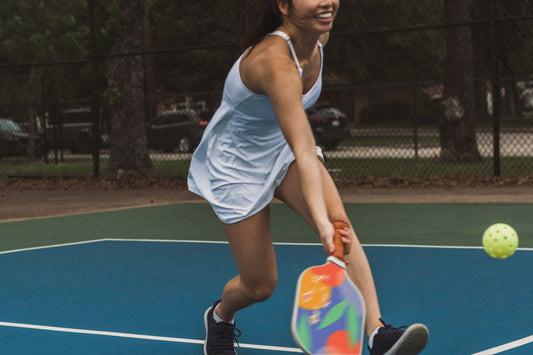 Girl swinging Court pickleball paddle at a pickleball with a white tennis outfit on.