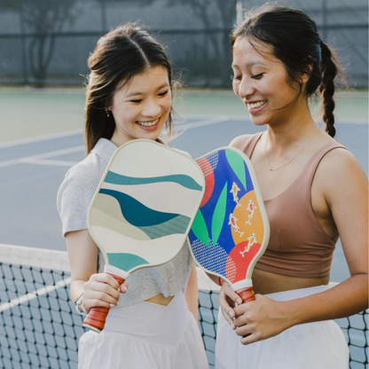 two girls smiling and holding Court pickleball paddles standing in front of a pickleball net